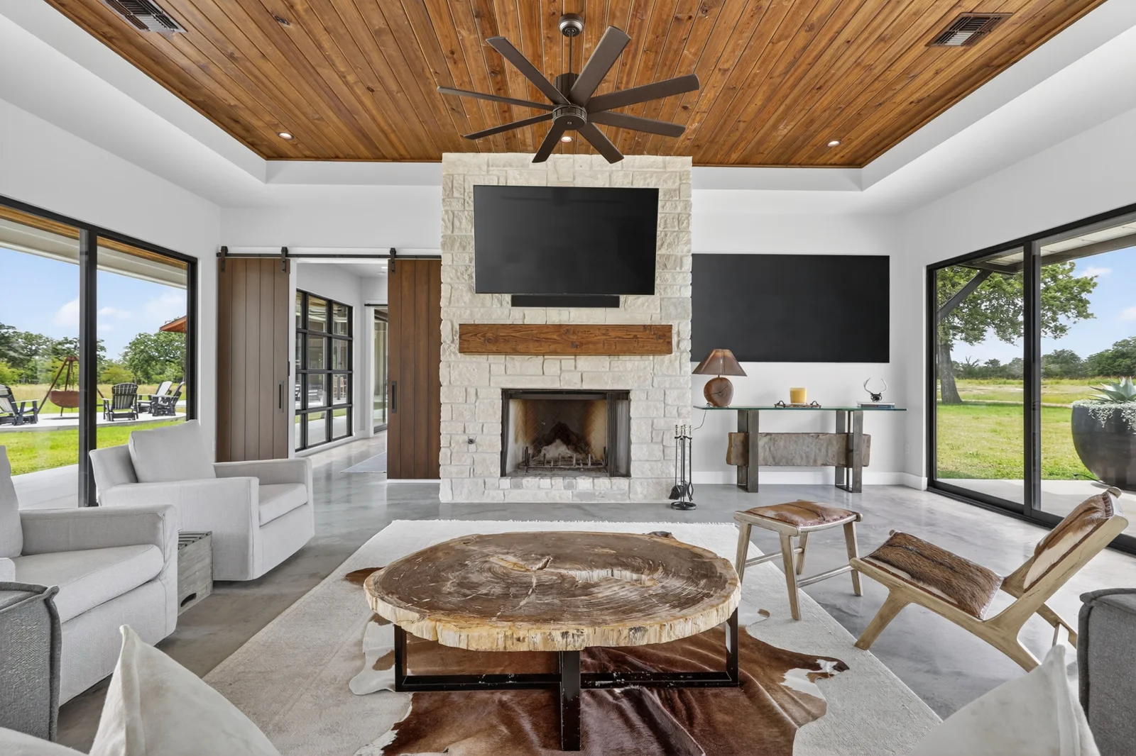 Living room with a wood ceiling, stone fireplace, and large windows overlooking the yard.